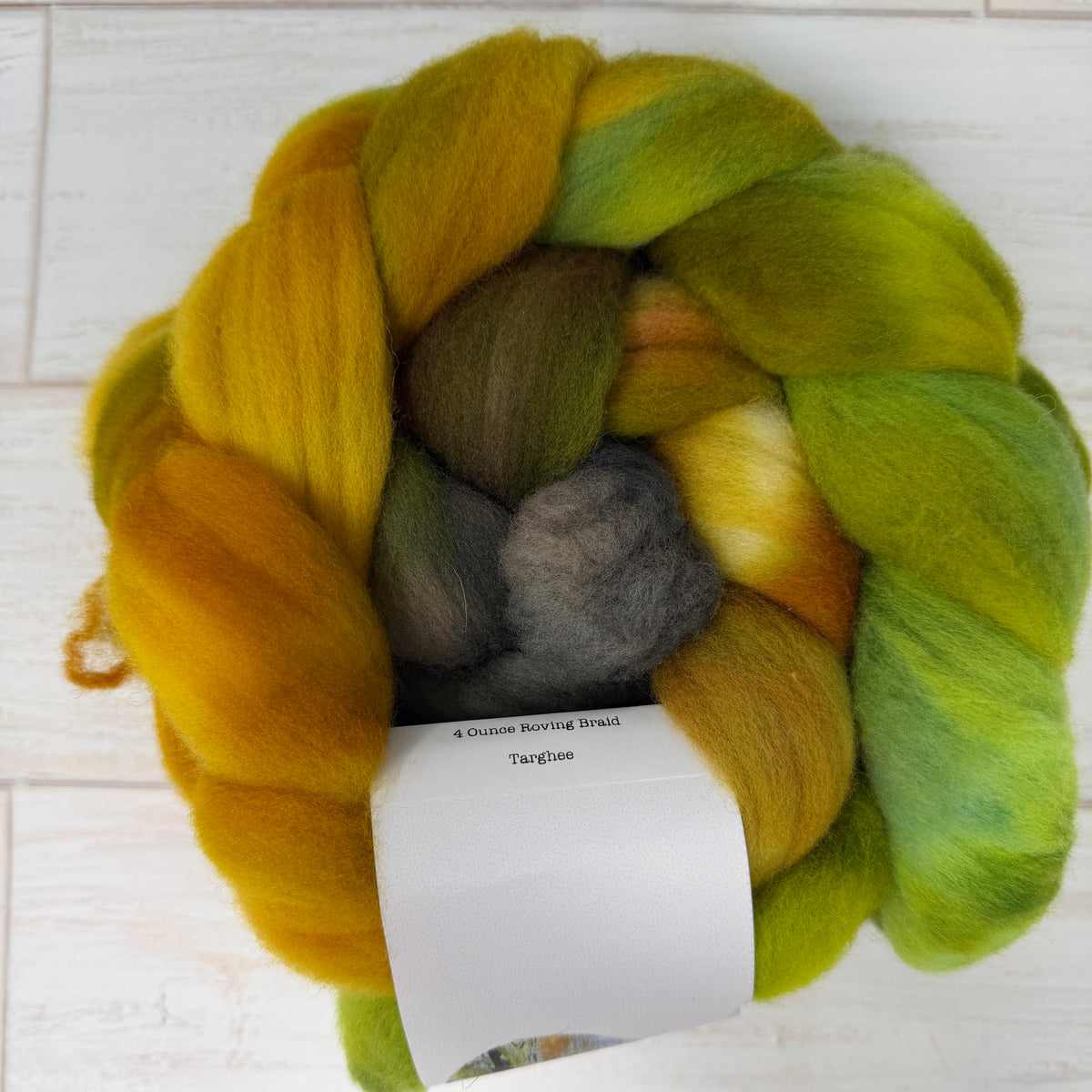 A hand-bundled hank of fluffy wool roving, shown from above on a light wood background. The fibers have a variegated color story of golden yellow, olive green, lime green, and a central gray section.