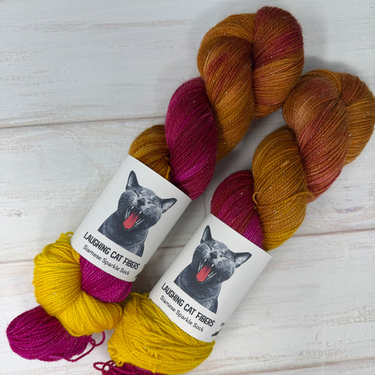 Three skeins of yarn in yellow, pink, and brown with 'Laughing Cat Fibres' labels on a light wooden surface.