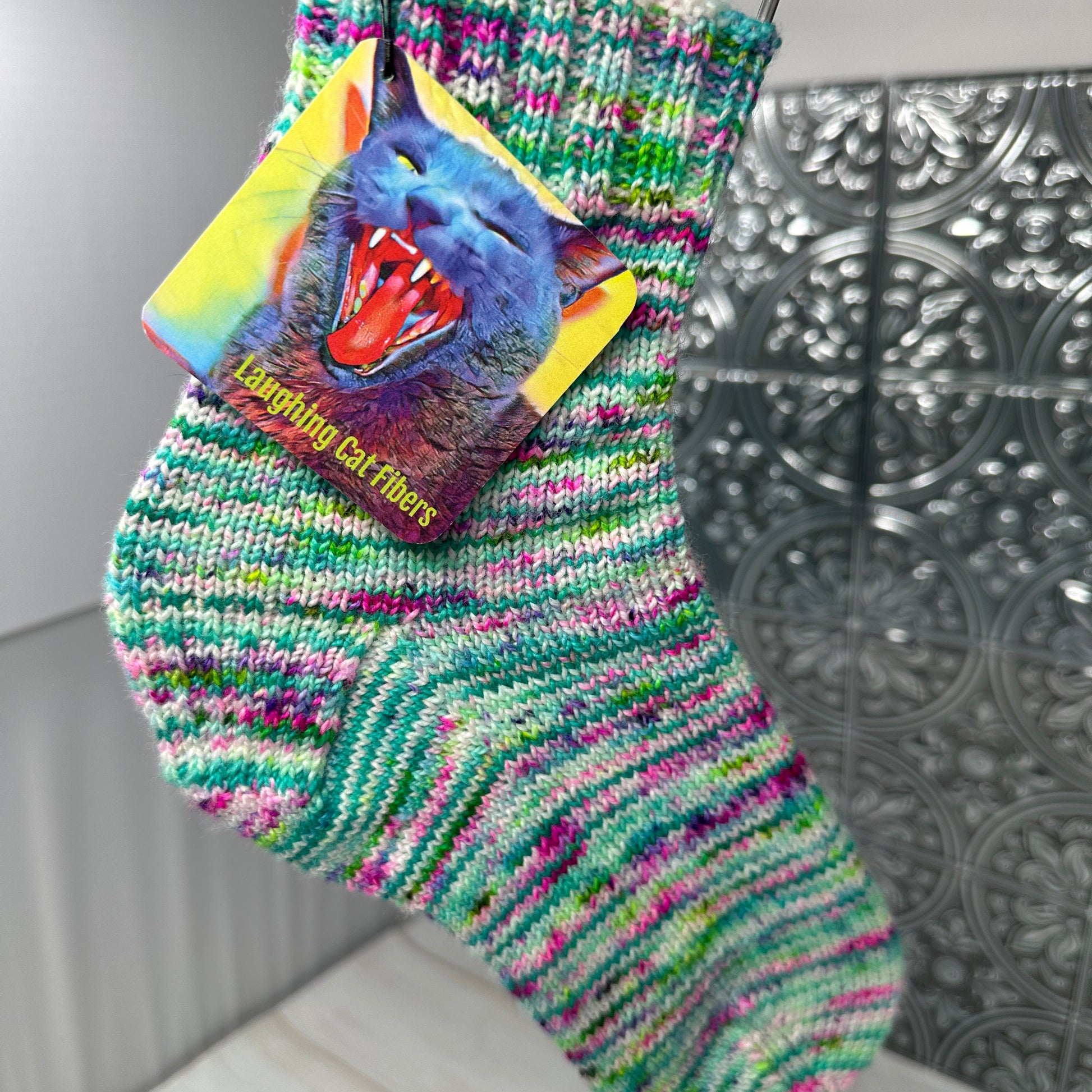 Colorful knitted sock with a cat design and brand tag hanging against a metallic wall.
