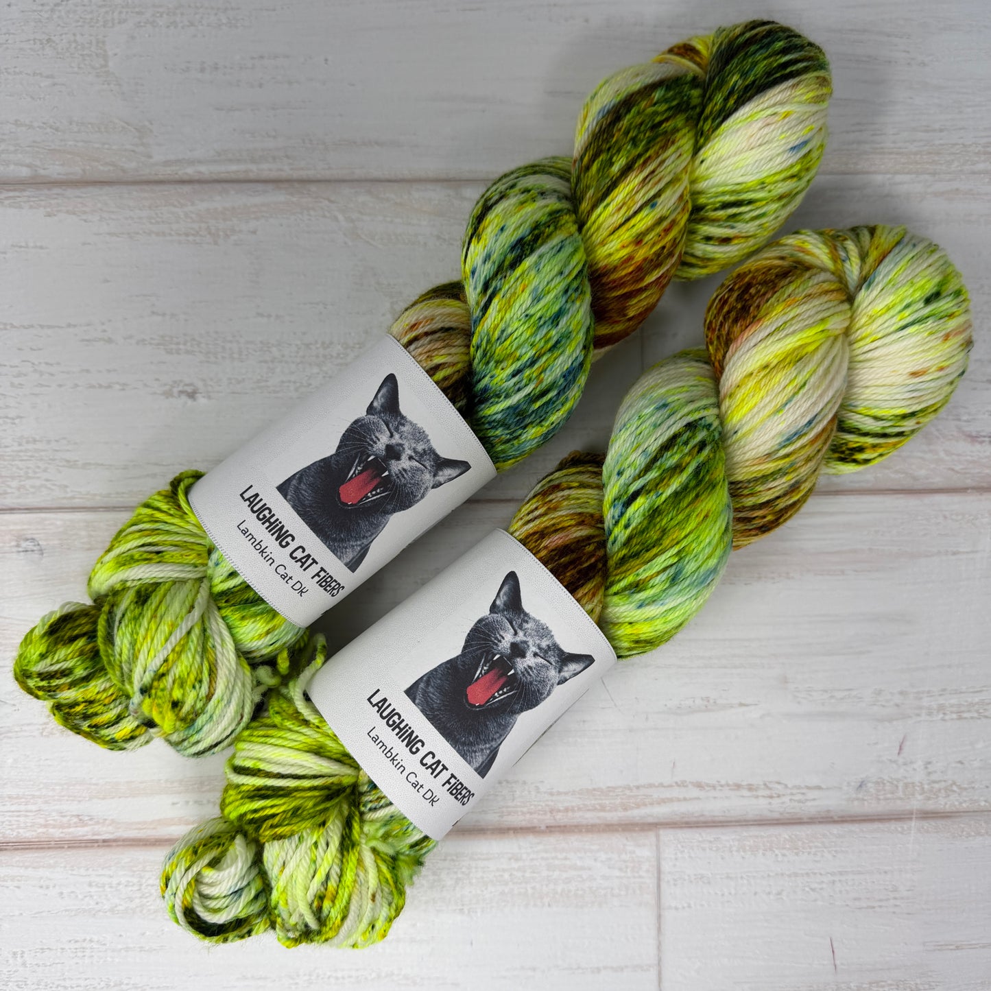 Two skeins of green and yellow yarn with 'Laughing Cat Fibres' labels on a light wooden surface.