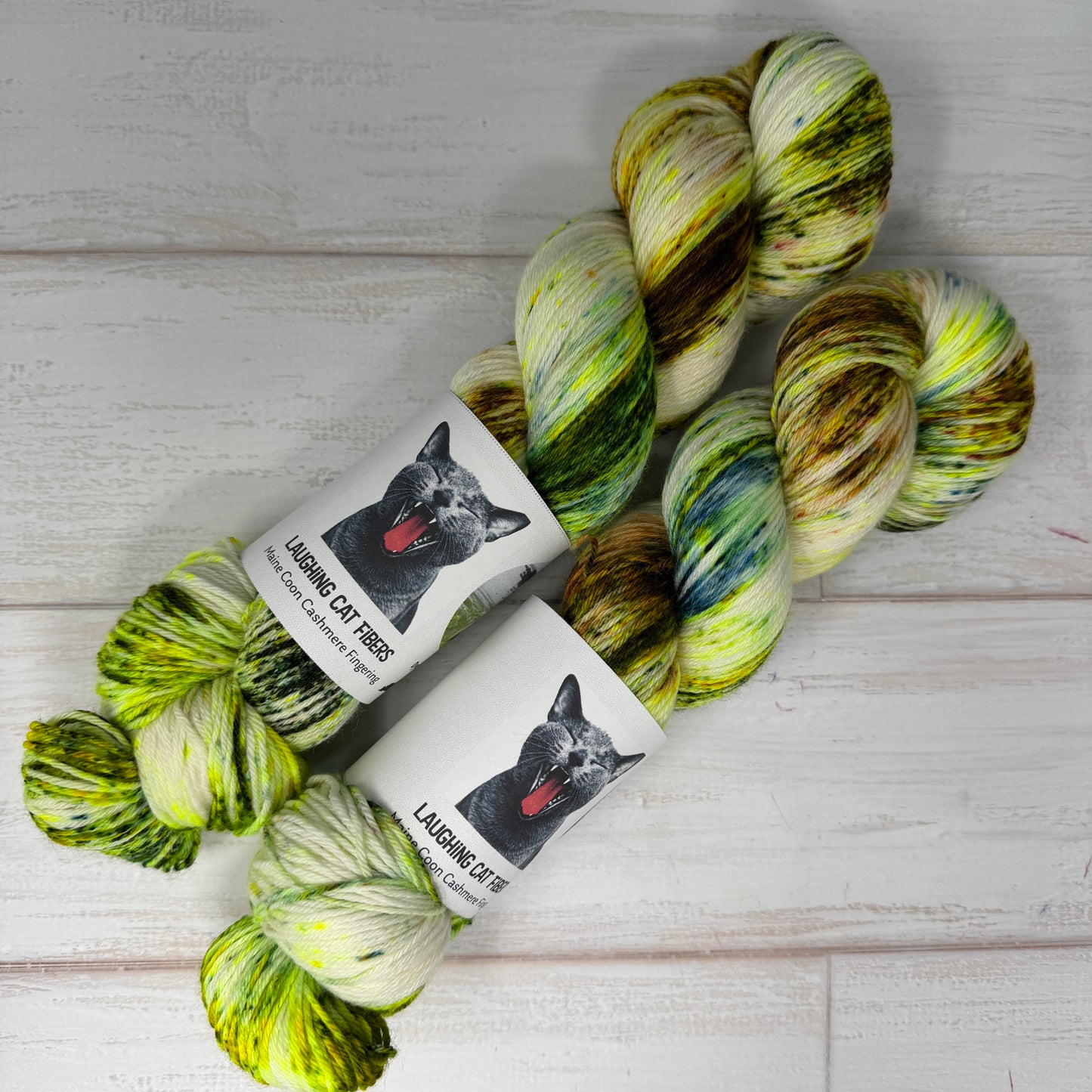Three skeins of multicolored yarn with a 'Laughing Cat Fibres' label on a wooden surface.