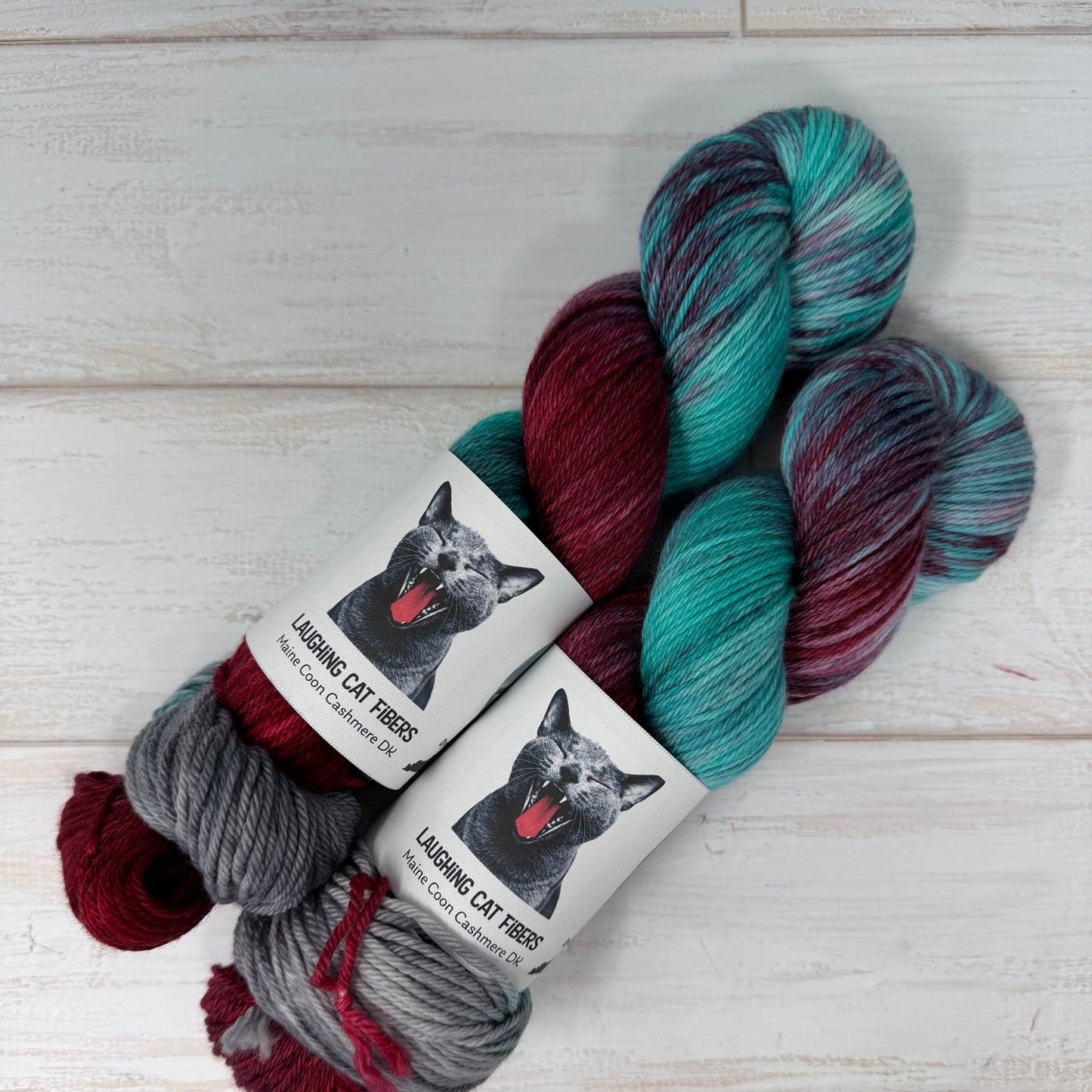 Three skeins of yarn in teal, maroon, and gray with labels on a light wooden background