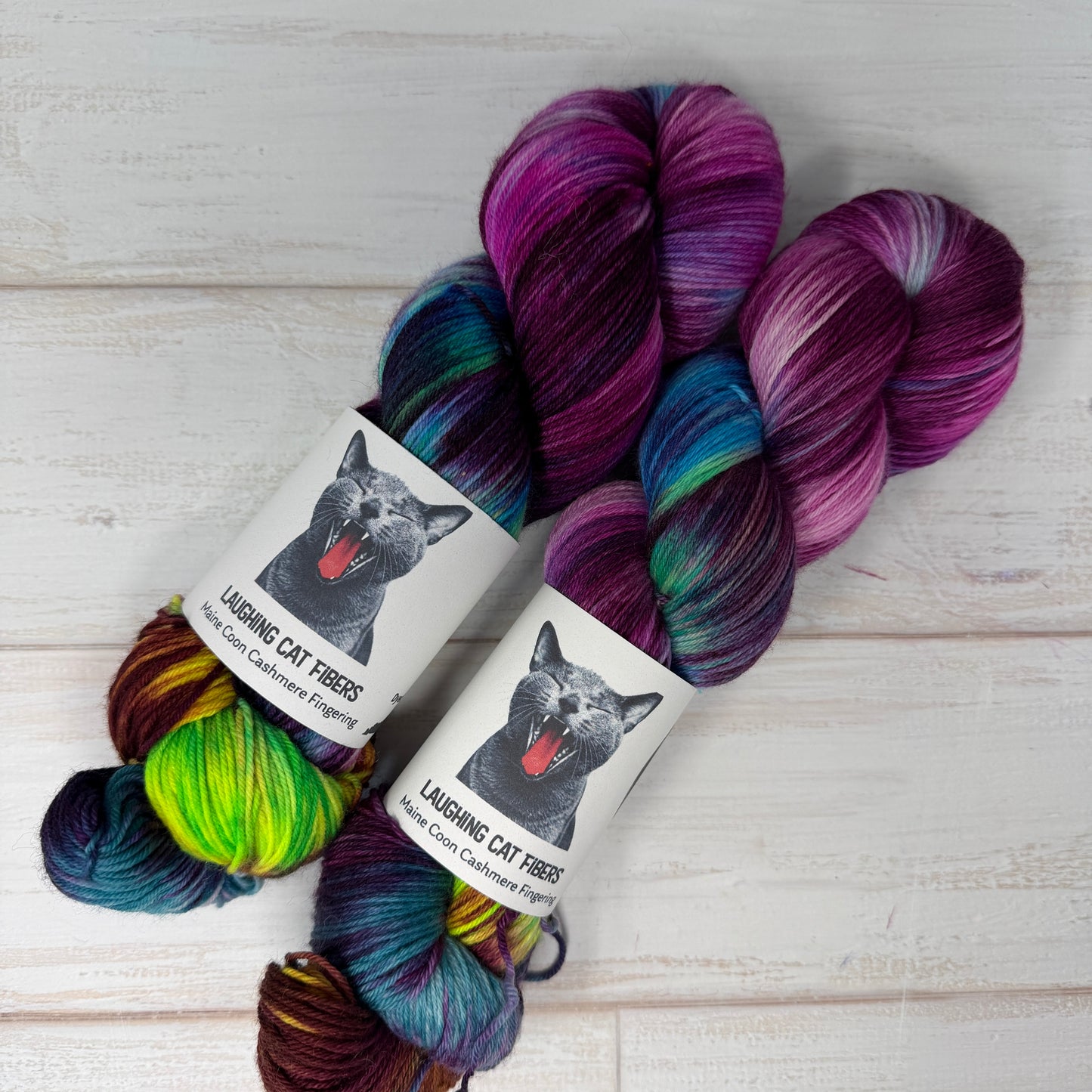 Two skeins of multicolored yarn with 'Laughing Cat Fibres' labels on a wooden surface.