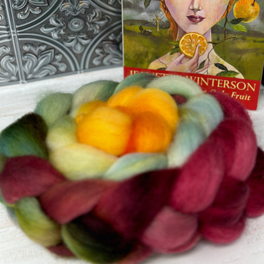 A braided wool roving in shades of orange, green, and purple, with a printed card showing a illustration of a person with orange hair and a book cover in the background.