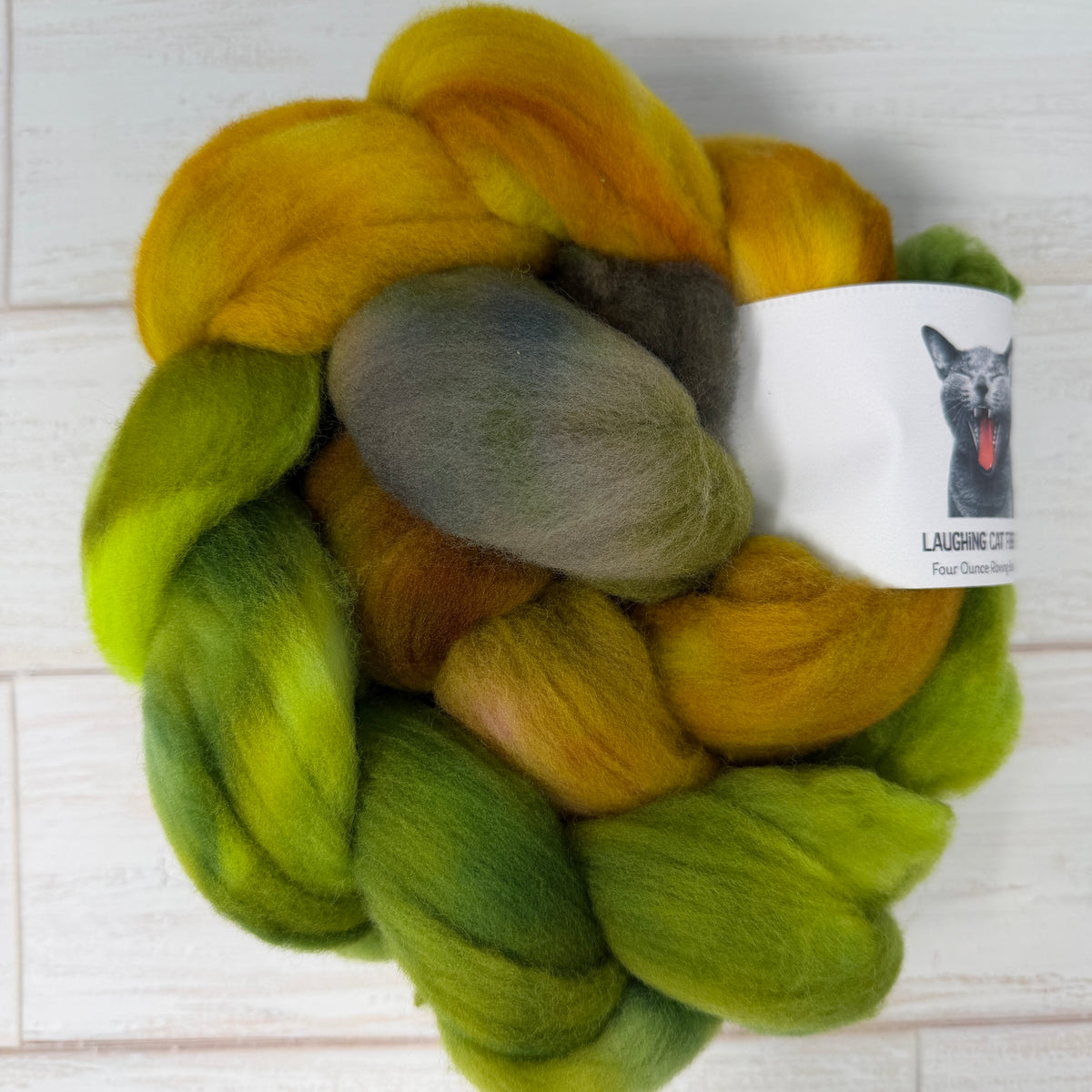 A ball of fluffy wool roving or fiber blend in a marbled colorway of yellow, green, and gray, with a white paper label attached. The label features a black dog illustration and the text 'LAUGHING LOT Four Duchies Farm'.
