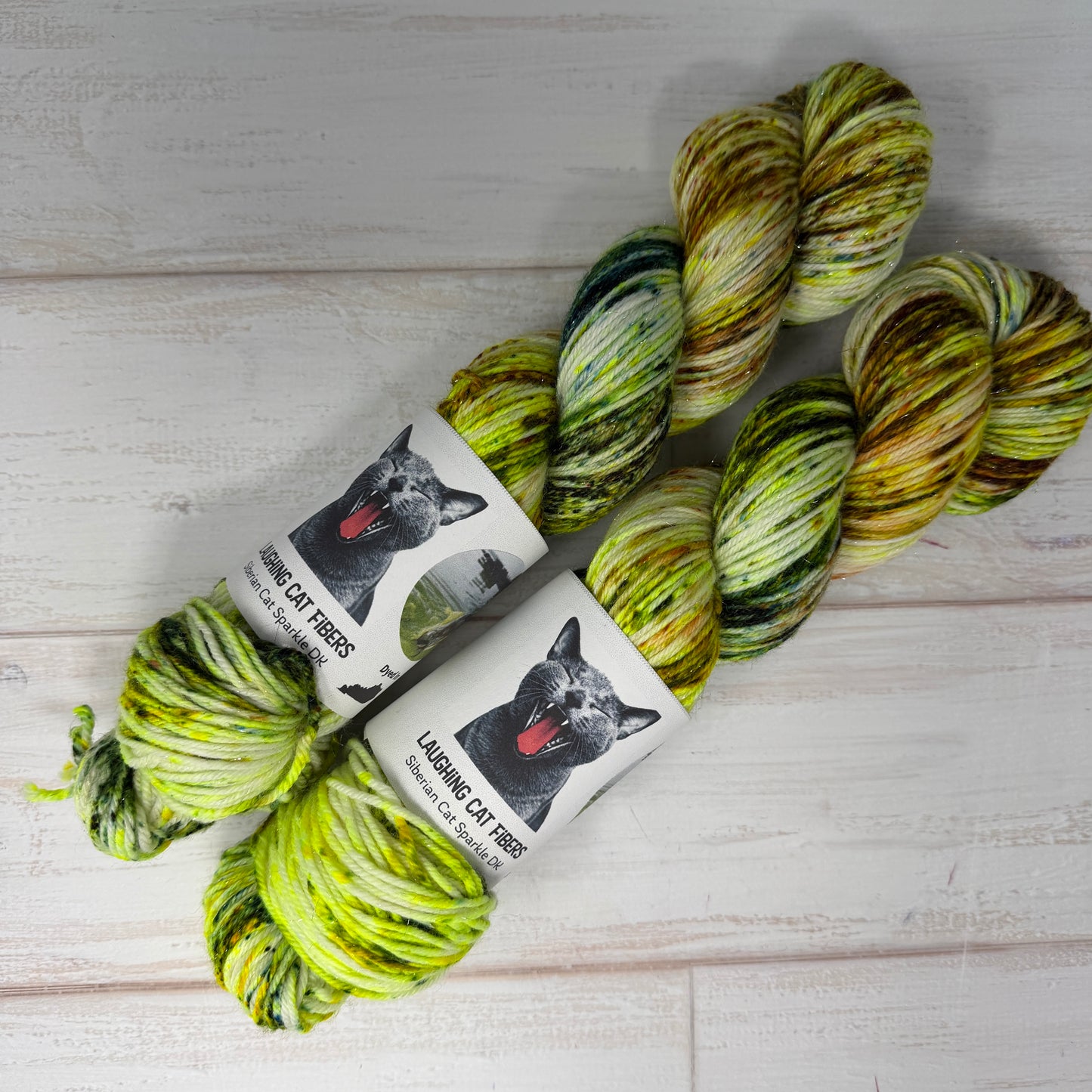 Two skeins of green and yellow yarn with 'Laughing Cat Fibres' labels on a light wooden surface.