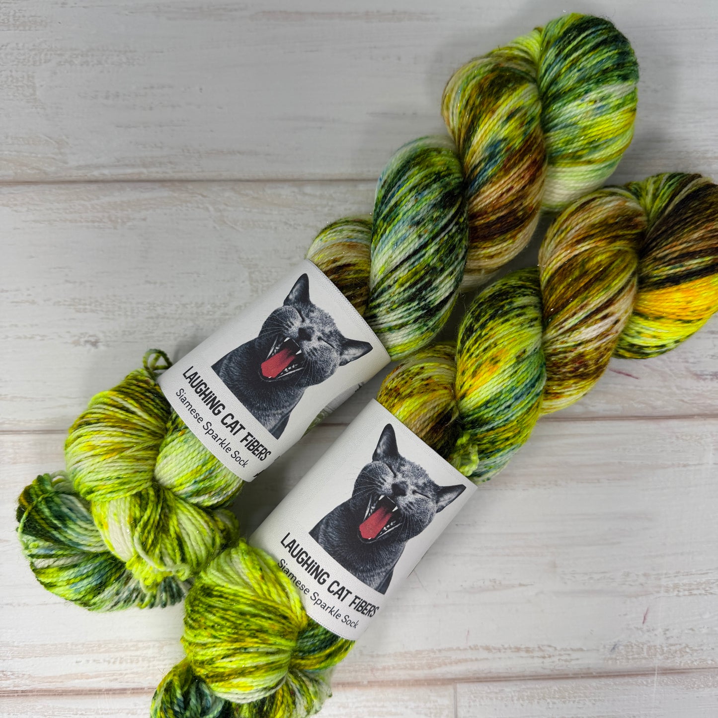 Two skeins of green and yellow yarn with 'Laughing Cat Fibres' labels on a light wooden surface.
