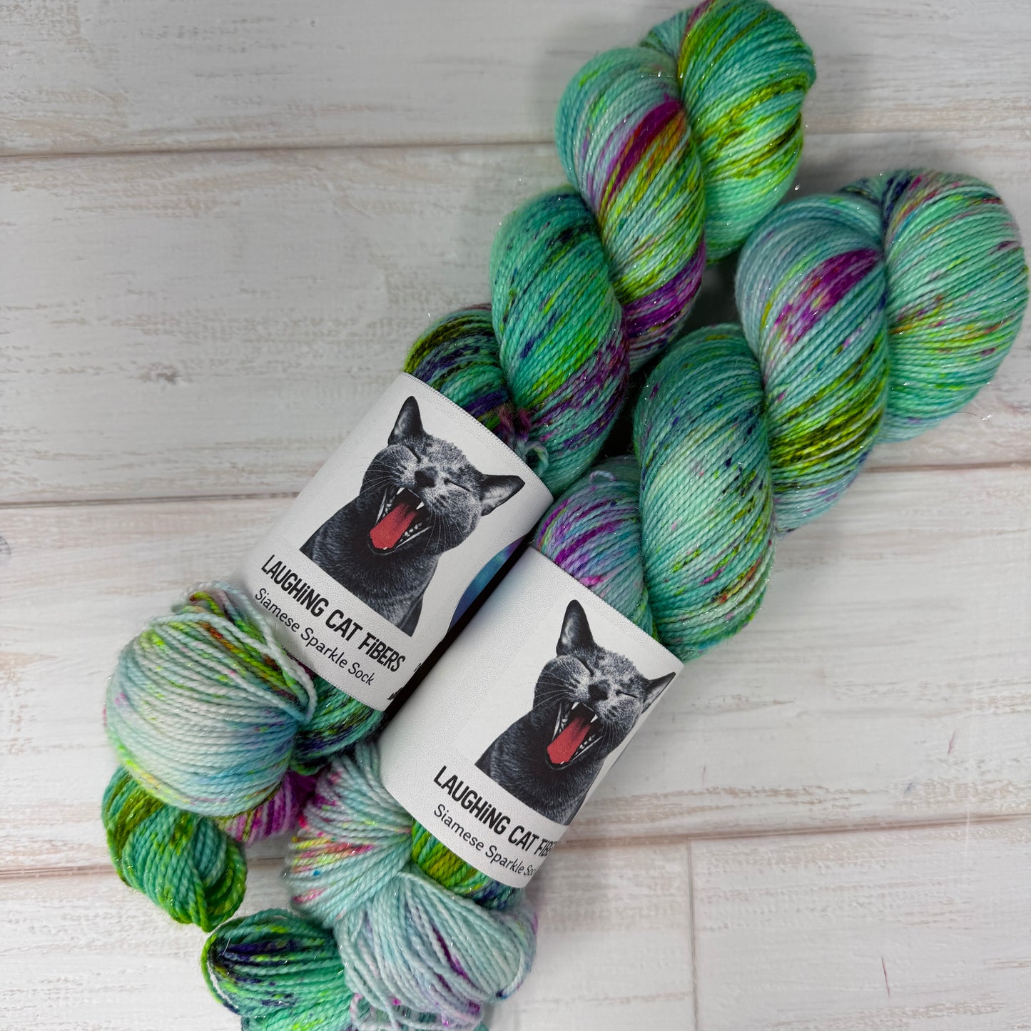 Two skeins of colorful yarn with 'Laughing Cat Fibres' labels on a wooden surface.
