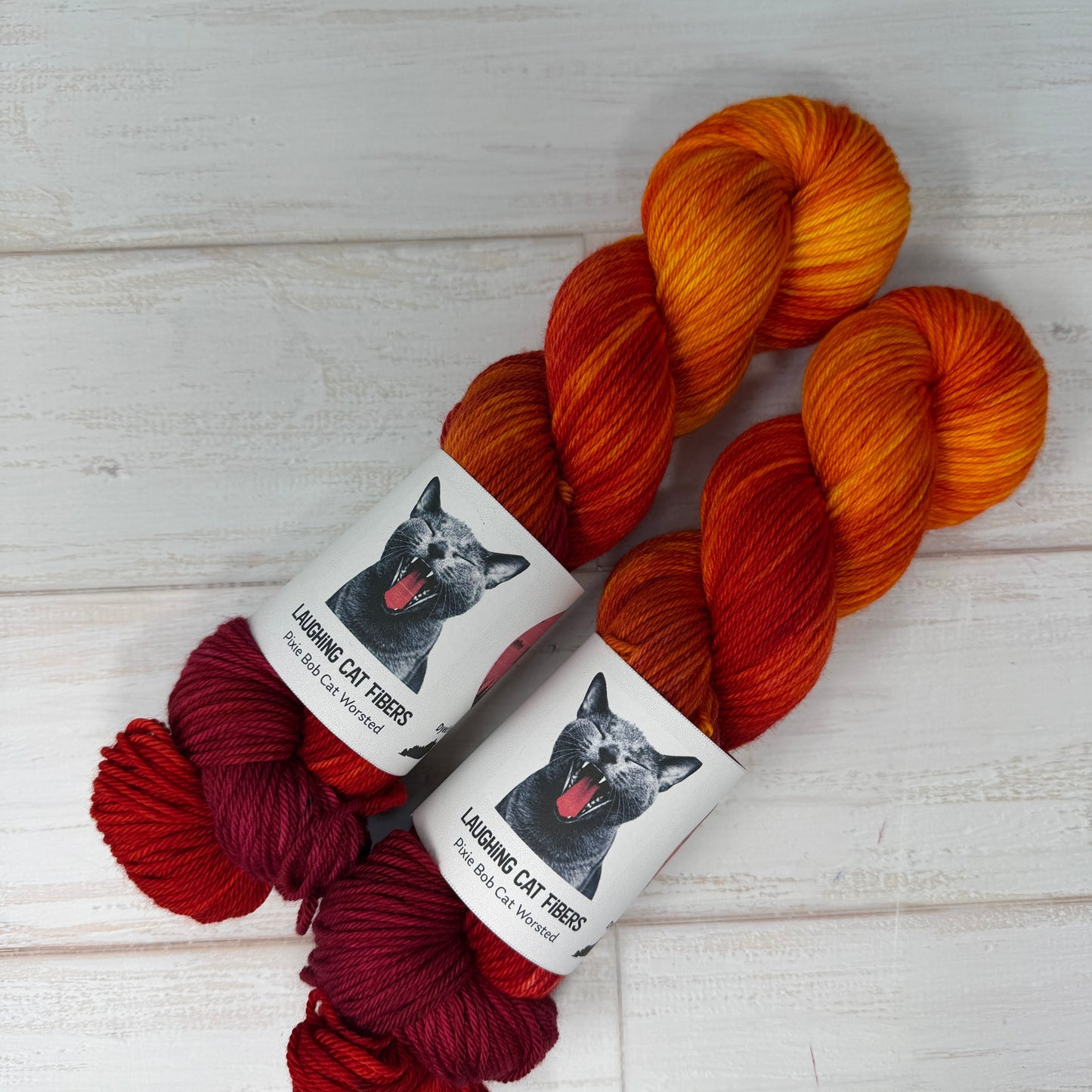 Two skeins of yarn in orange, red, and burgundy with a label on a light wooden surface.