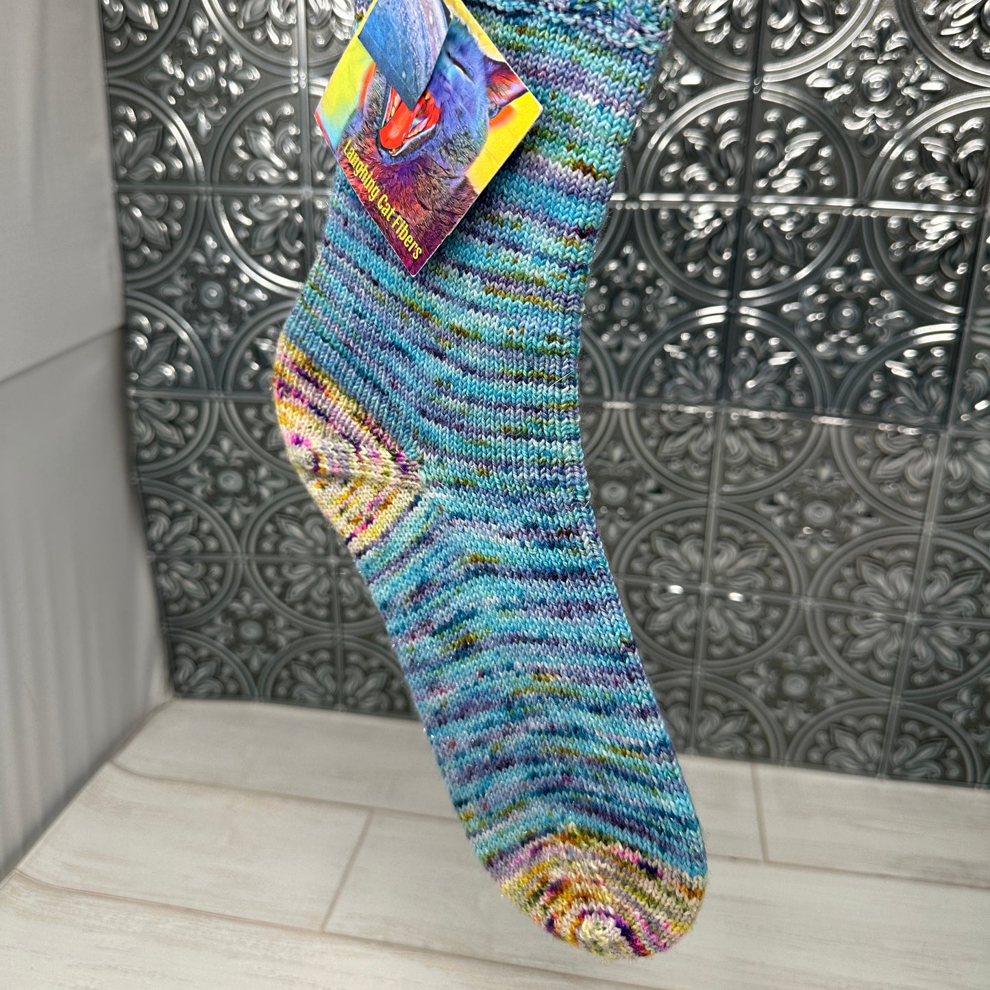 Colorful sock with a brand tag hanging against a tiled wall.