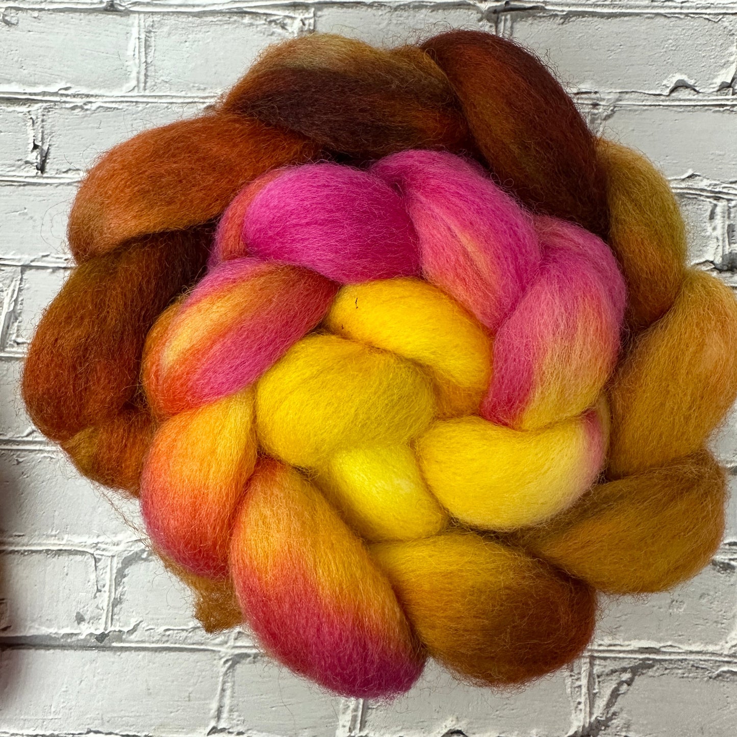 A braid of wool roving dyed in shades of blue, pink, yellow, and orange, displayed against a brick wall background.